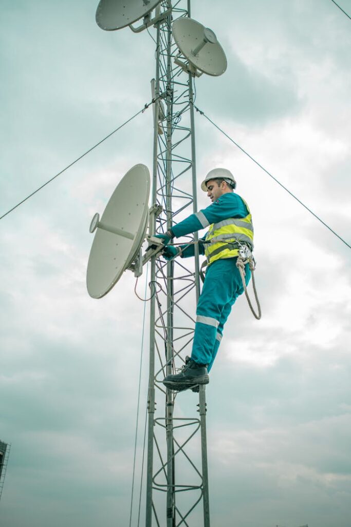 A worker in safety gear installs a satellite dish on a telecom tower under an overcast sky.